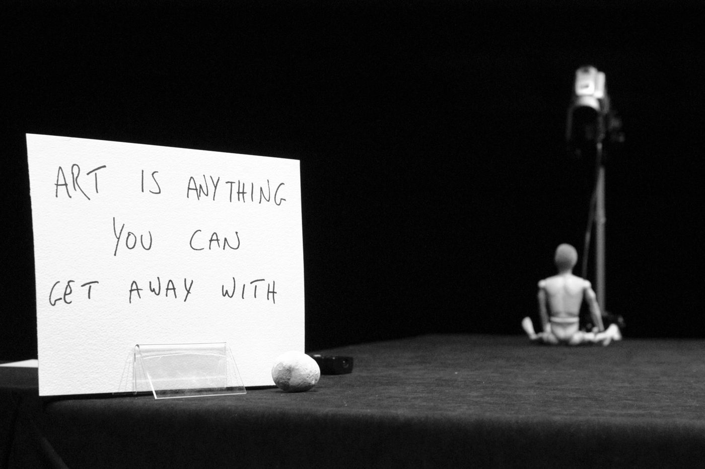 A paper sign stating 'ART IS ANYTHING YOU CAN GET AWAY WITH' with a doll and camera in the background.