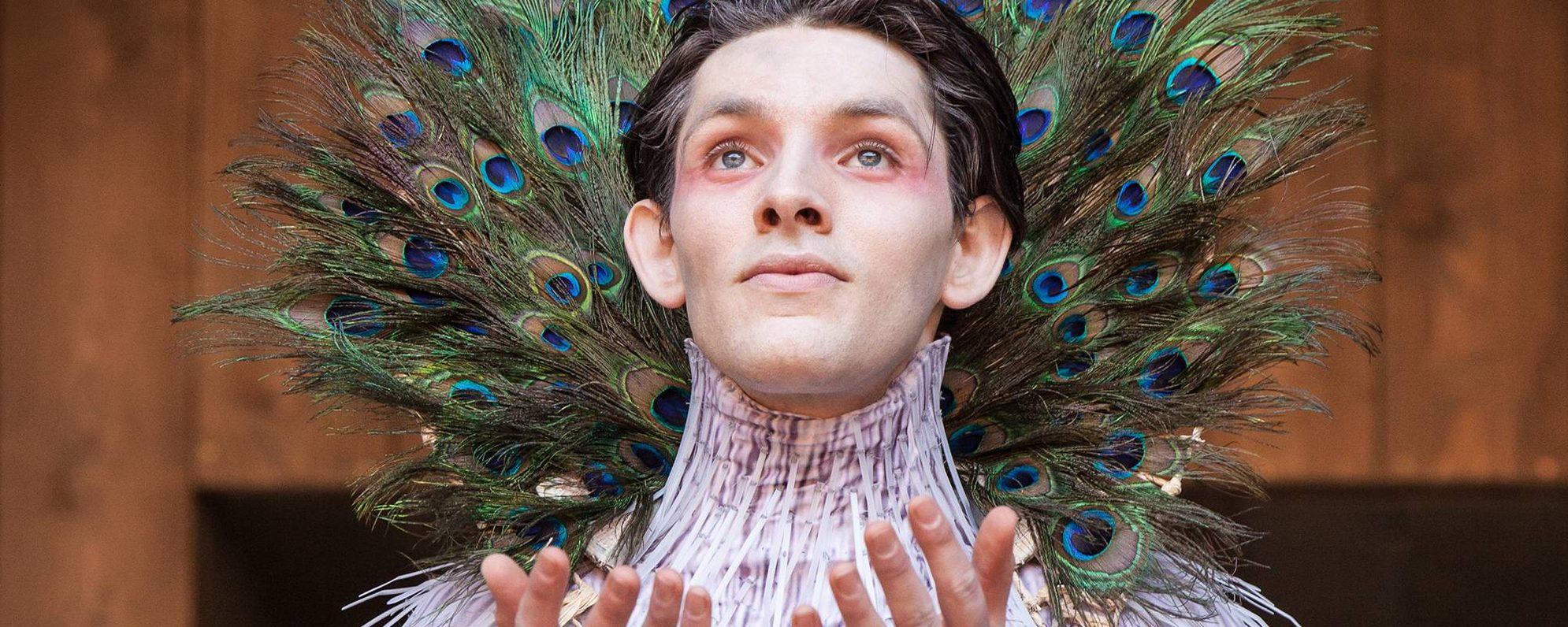 An actor wearing a pink feathered costume and eye makeup, with peacock feathers fanned out in a plumage around their neck, holds out their hands palms upwards, looking up.