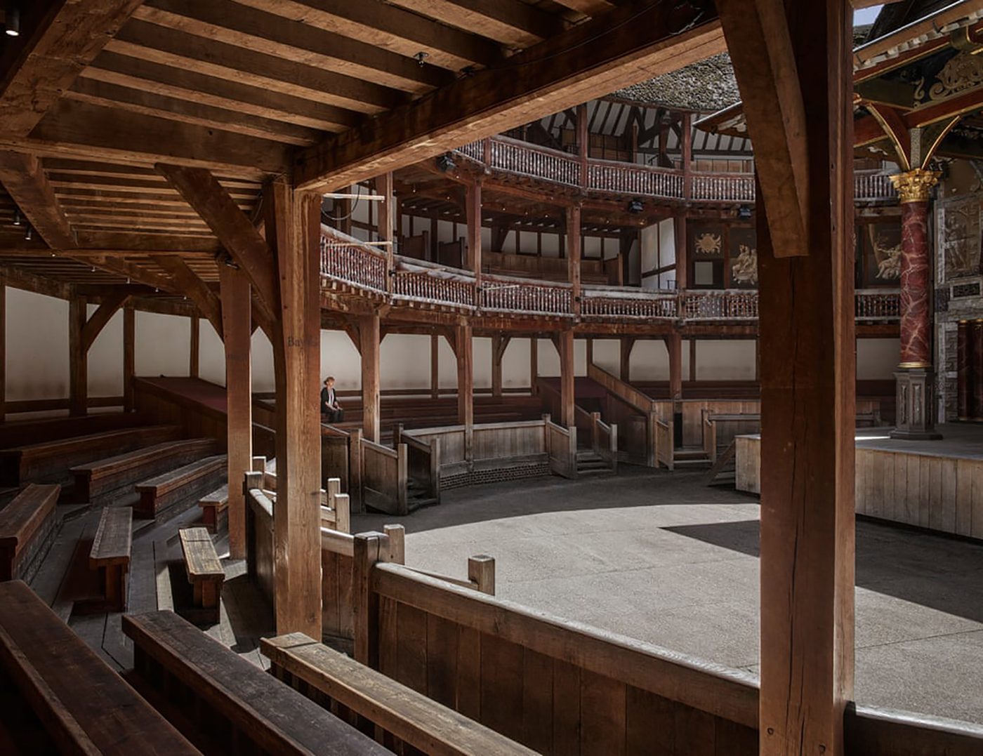 A woman sits in the lower gallery of a circular timber theatre, which lays empty.