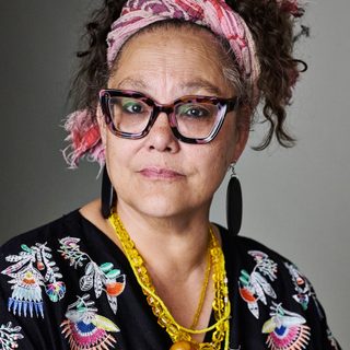 Naomi Wirthner looking directly at the camera. Naomi is wearing a patterned shirt, necklaces, and a pink cloth headband.