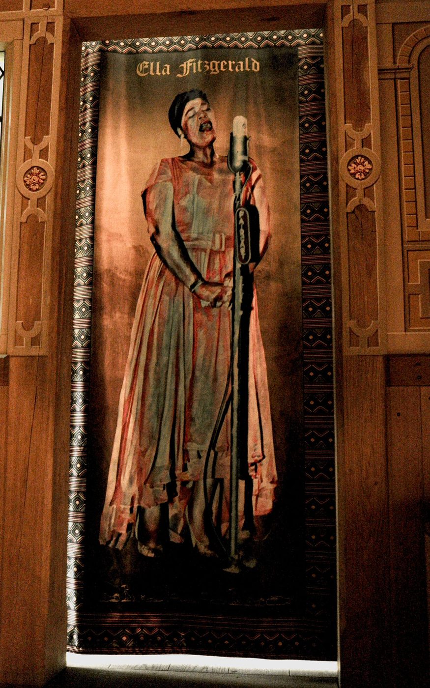 A tapestry of a person standing and singing into a microphone, wearing a long pink dress, hangs within a wooden door frame. Around the border of the tapestry is a patterned fabric.
