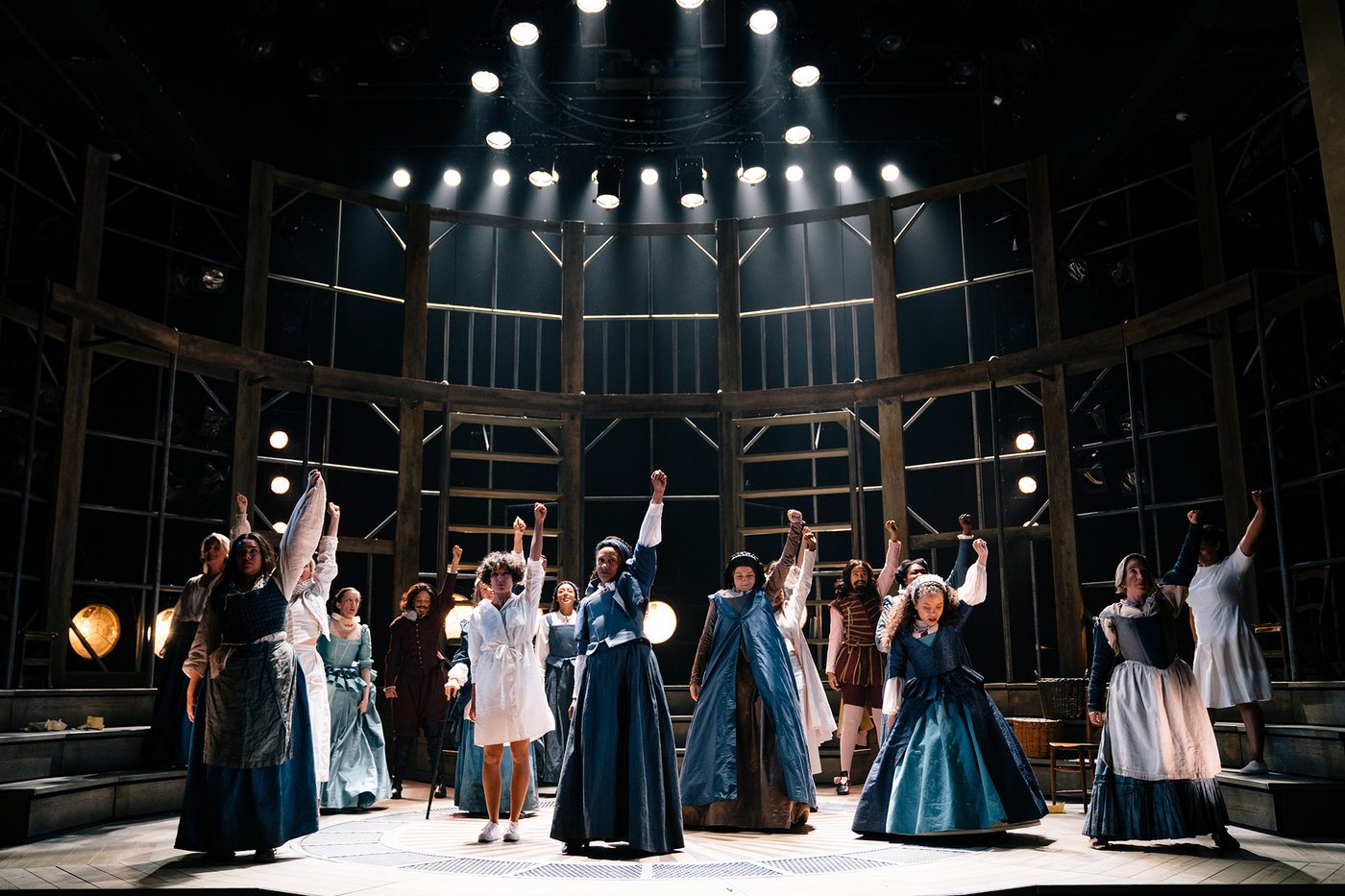 A group of actors stand on stage under spotlights, their left arms raised in the air into a fist.