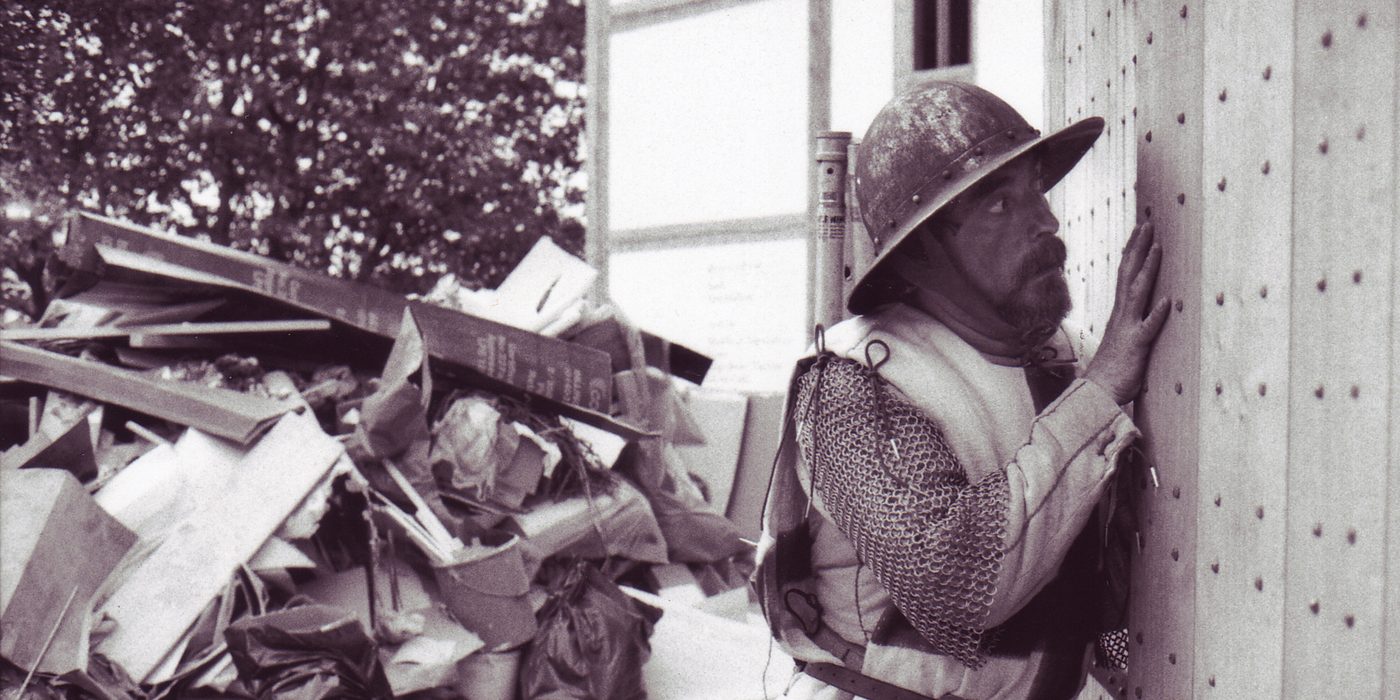 An actor wearing armour leans his ear against the wooden oak doors of a timber theatre.