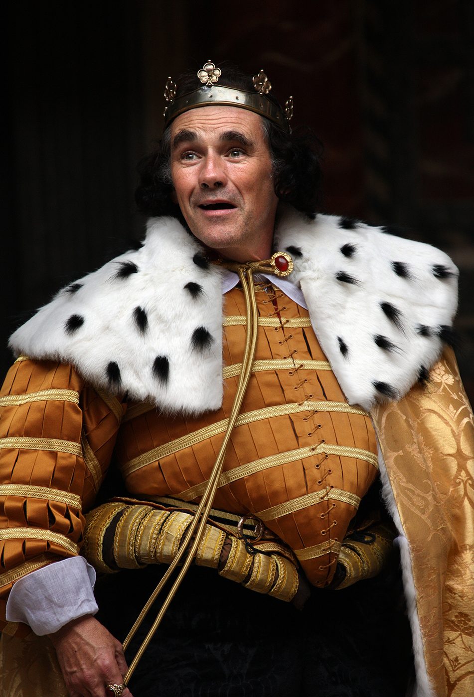 A man wearing an ermine royal cloak and golden doublet smiles.