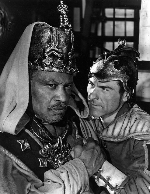 A black and white photograph of two actors, one leans in close, menacingly, towards the other.
