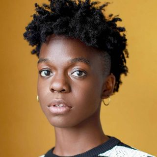 Ryn Alleyne looking directly at the camera. They have short black hair and are wearing a striped black and white top, in front of a yellow background.