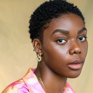 Selina Jones looking directly at the camera. They have short black hair and are wearing gold jewellery and a pink and orange shirt.