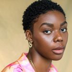Selina Jones looking directly at the camera. They have short black hair and are wearing gold jewellery and a pink and orange shirt.