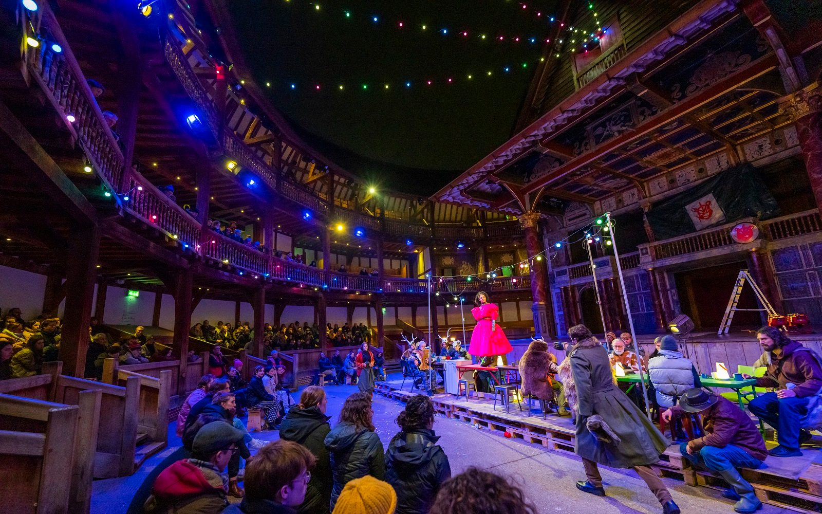 A large group of actors are performing in the Yard of the Globe Theatre at night, with seated audience members watching them.