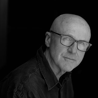 Black and white image of Tim Crouch wearing a dark shirt and looking sideways towards the camera.