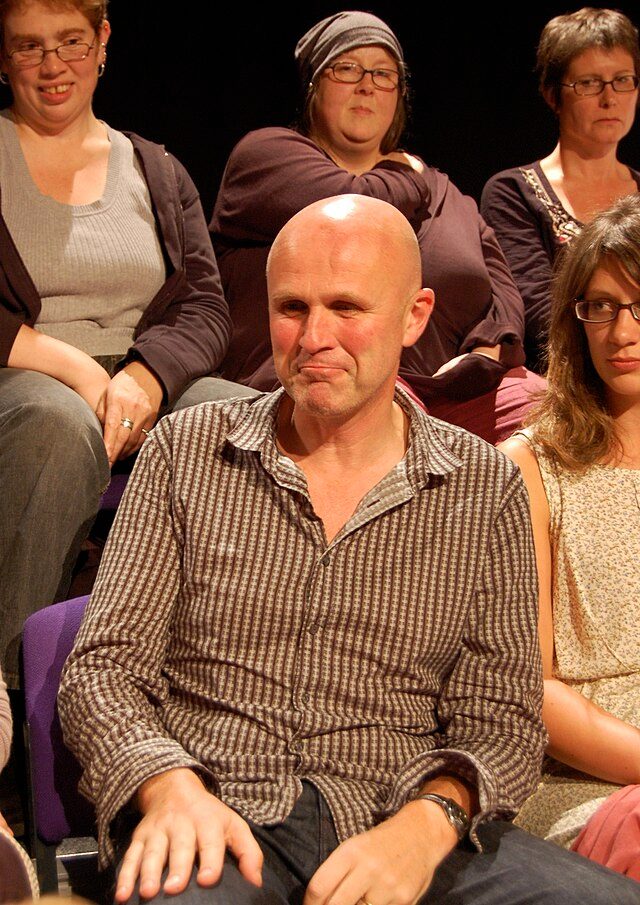 Actor Tim Crouch wears a black and white dotted shirt sitting in a chair surrounded by audience members.