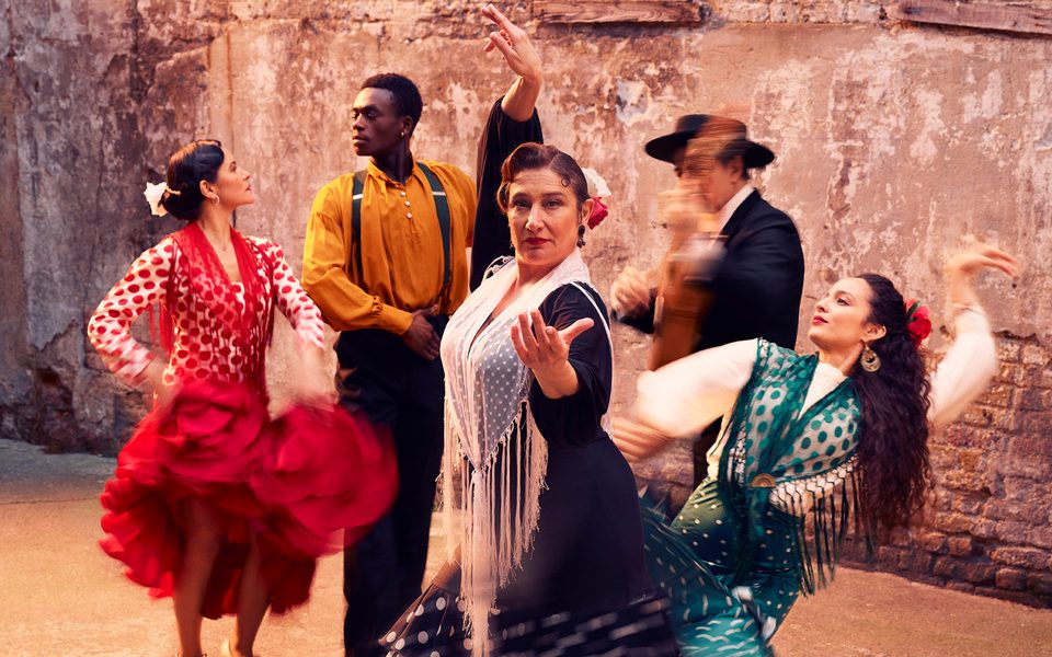 A group of flamenco performers are captured in motion against a plain brick wall. Four of the group, wearing traditional colourful flamenco costumes, take strong dance positions. A fifth person wearing a dark suit stands playing a guitar, which is blurred through the capture of movement.