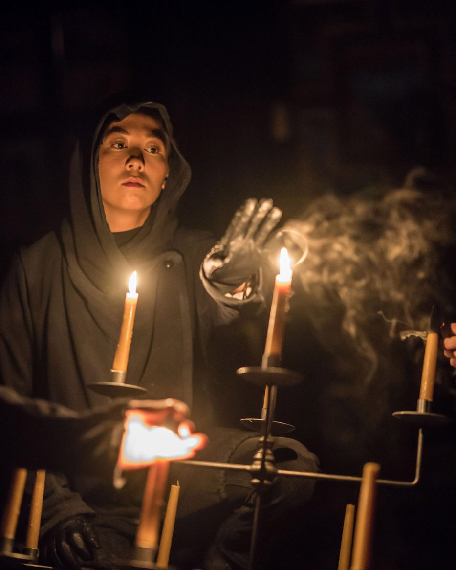 A cloaked man raises his hand to a lit candle.