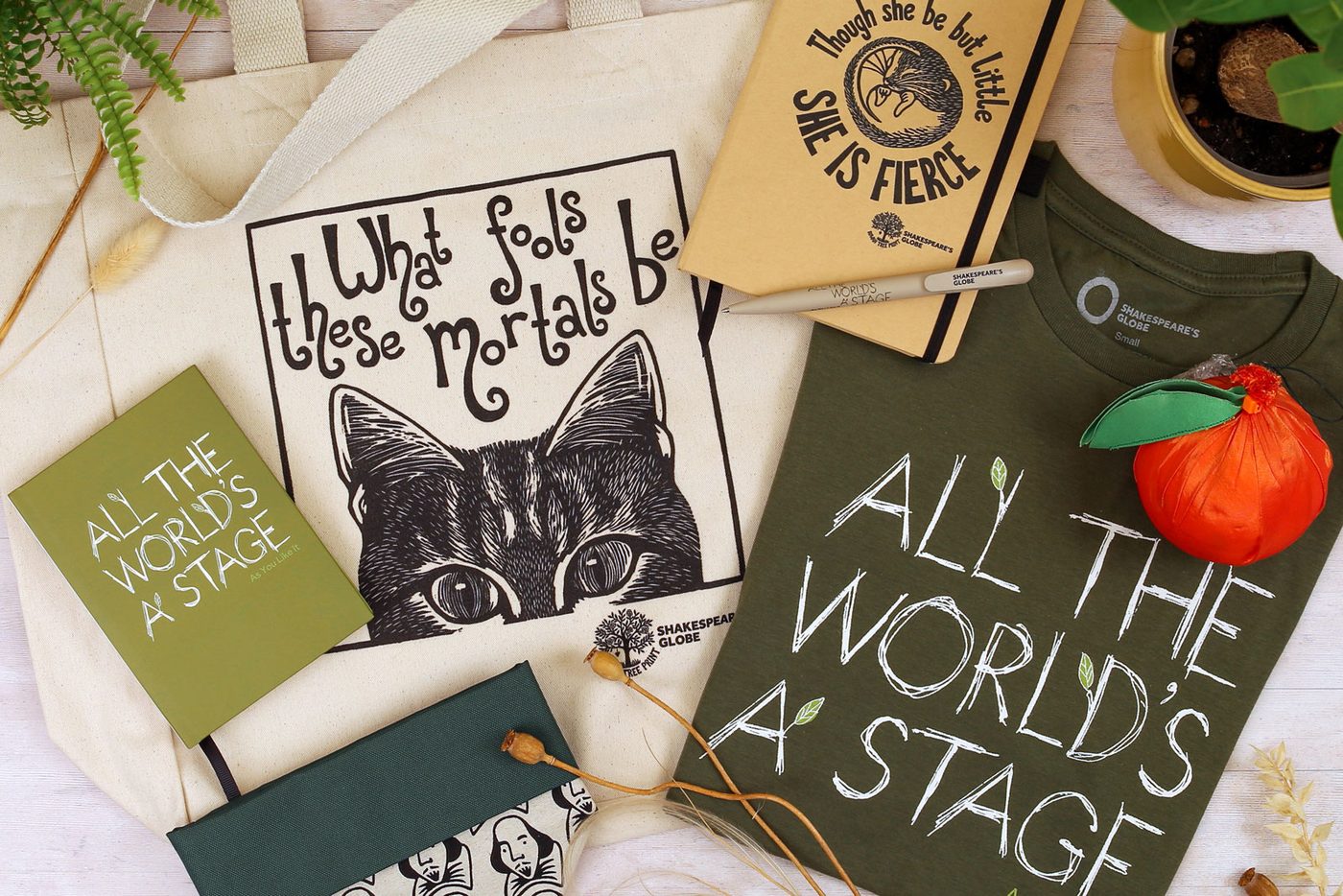 Shakespeare merch including a tote bag, notebooks, t-shirt, pen, and plant holder are displayed on a white wood background surrounded by plants.