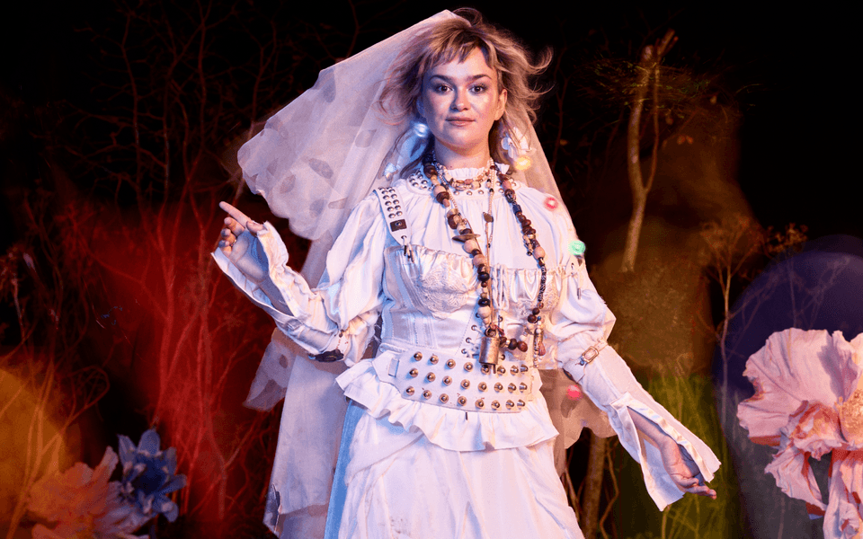 A person wearing a white shirt and a pinafore dress with a veil, smiles gently directly into the camera, whilst pointing up with their right hand. They have blonde hair and brown eyes and wear a large bead necklace in various shades of brown. Small orbs of colour appear around them. In the background, various trees are overshadowed by tints of red, yellow and purple.